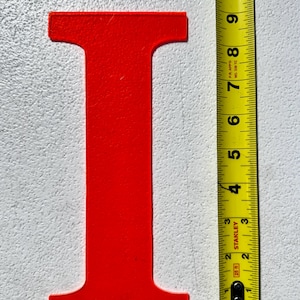 May include: A bright red letter "I" is positioned next to a yellow and black measuring tape. The letter is approximately 7 inches tall. The measuring tape shows inches and feet.