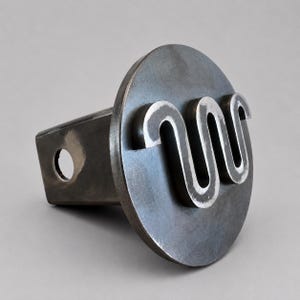 May include: A dark gray metal trailer hitch cover featuring a circular face plate with a raised, stylized, continuous wavy line design. The square-shaped mounting shank extends from the back and includes a circular hole for a hitch pin.
