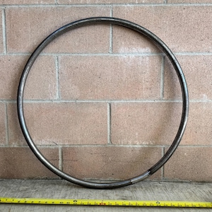 May include: A circular metal hoop, approximately 30 inches in diameter, rests against a brick wall. The hoop is made of a dark, possibly steel, material. A yellow measuring tape is positioned below the hoop.