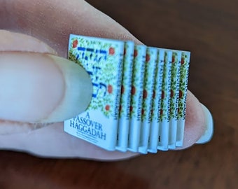 May include: A stack of miniature Passover Haggadah books with a white background and green and red floral designs. The top book has the text "A Passover Haggadah" printed on the front.