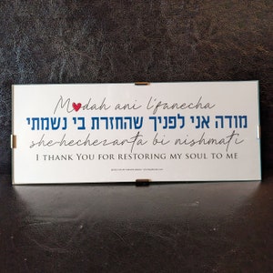 May include: A framed white sign with a blue and red heart. The sign has the text 'Modah ani l'fanecha' in cursive script, followed by the Hebrew text 'מודה אני לפניך שהחזרת בי נשמתי' and the English text 'she-hechezarta bi nishmati' and 'I THANK YOU FOR RESTORING MY SOUL TO ME'.