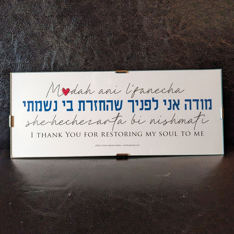 May include: A framed white sign with a blue and red heart. The sign has the text 'Modah ani l'fanecha' in cursive script, followed by the Hebrew text 'מודה אני לפניך שהחזרת בי נשמתי' and the English text 'she-hechezarta bi nishmati' and 'I THANK YOU FOR RESTORING MY SOUL TO ME'.