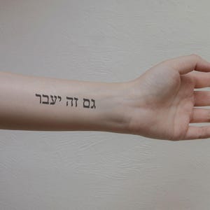 May include: A temporary tattoo on a forearm with the Hebrew phrase "Gam ze ya'avor" in black ink. The phrase translates to "This too shall pass". The skin tone is light, and the background is a textured, off-white surface.