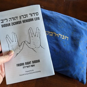 May include: A white booklet with the title "Siddur Zichron Yehuda Leib" and the text "Friday Night Siddur" in black lettering. The booklet features two hand drawings of the Vulcan salute, a popular gesture from the Star Trek franchise. The booklet is lying on a blue fabric with gold Hebrew lettering.