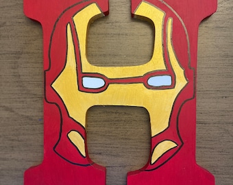 May include: A wooden letter "H" painted in red, with a gold and black design resembling Iron Man's mask. The eyes are white. The letter is likely for decoration or a themed room.