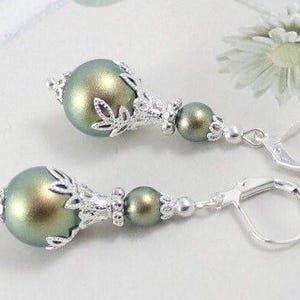May include: Dangle earrings with iridescent green-gold beads and silver-tone metal detailing. The earrings have a lever-back clasp and a floral design on the top bead. The earrings are presented on a white surface.