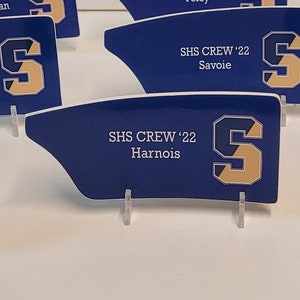 May include: Ten blue and gold rowing team nameplates with the text "SHS CREW '22" and the name of each crew member. The nameplates are on small clear acrylic stands.