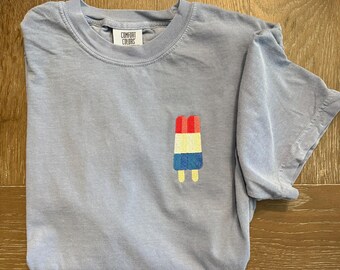 May include: A light blue t-shirt with a small embroidered popsicle design in red, white, blue, and yellow. The shirt has a crew neck and short sleeves. The brand name "COMFORT COLORS" is on a tag near the collar.