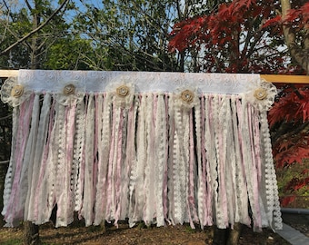 May include: A white and pink lace curtain with burlap flowers and a wooden dowel rod. The curtain is made of various lengths of lace and ribbon.