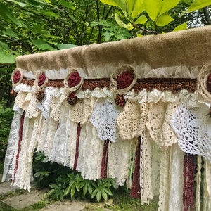 以下が含まれることがあります： 白とクリーム色のレースと、ワインレッドの布の花が並んだ、素朴なジュートのバルーン。バルーンは、白とクリーム色のレースとワインレッドの布のフリンジが付いています。