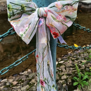 Puede incluir: Un lazo blanco de gasa con flores bordadas en tonos de rosa, rojo, verde y amarillo. El lazo está atado alrededor de un poste de metal oxidado.
