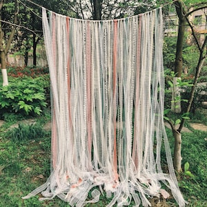 May include: A decorative wall hanging made of long, flowing fabric strips in white, pink, and green. The strips are suspended from a horizontal line, creating a textured, bohemian-style backdrop. The backdrop is outdoors, surrounded by greenery.