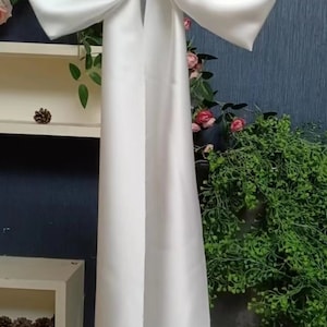 Op de afbeelding: Een witte satijnen strik met lange linten, hangend aan een houten stok. De strik is geknoopt in een grote, elegante knoop.
