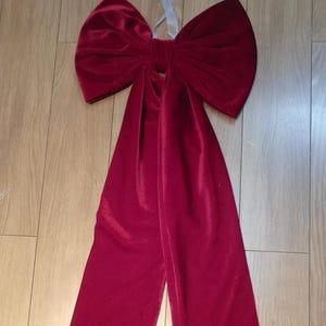 Puede incluir: Un gran lazo de terciopelo burdeos con largas colas fluidas. Se adjunta una cinta plateada transparente para colgarlo. El lazo es probablemente para la decoración navideña o de regalo.