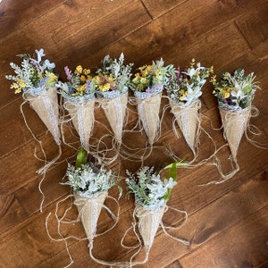 May include: Ten small bouquets of white and yellow flowers arranged in burlap cones with lace trim. The cones are tied with twine and the bouquets are arranged on a wooden surface.