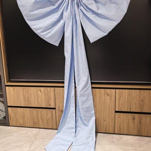 May include: A large, light blue fabric bow with long, flowing tails. The bow is suspended from a white string, and the fabric appears to be a lightweight material. The bow is against a dark background.