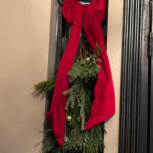 May include: A large, vibrant red velvet bow attached to a cluster of green evergreen branches. The arrangement includes small, round, metallic gold and green ornaments hanging from the foliage, creating a decorative seasonal display.