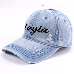 May include: Light blue denim baseball cap with distressed details. The word "Layia" is written in black script across the front. The cap has a curved peak and adjustable strap.