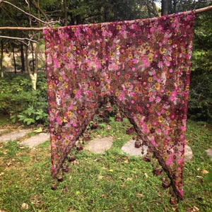 May include: A sheer, dark brown mesh fabric panel featuring an intricate floral embroidery pattern in shades of magenta, pink, and gold. The bottom edge is cut into a V-shape and is finished with a dark brown decorative trim and hanging tassels.