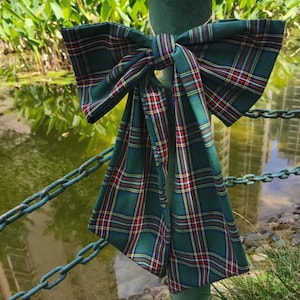 May include: A green, red, and yellow plaid bow tied around a green metal post. The bow is made of fabric and has a large, loose knot.