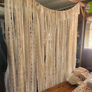 May include: A hanging curtain made of numerous vertical strips of cream and off-white lace, fabric, and ribbon. The strips are tied to a horizontal wooden rod, creating a layered, textured fringe effect that hangs down in long, flowing strands.