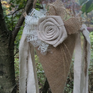 Puede incluir: Un cono de arpillera con un borde de encaje y una rosa de arpillera. El cono está colgado de una rama de árbol con una cinta blanca.