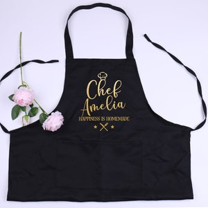 May include: Black apron with a gold inscription that reads "Chef Amelia Happiness is Homemade" with a chef's hat icon and crossed utensils.