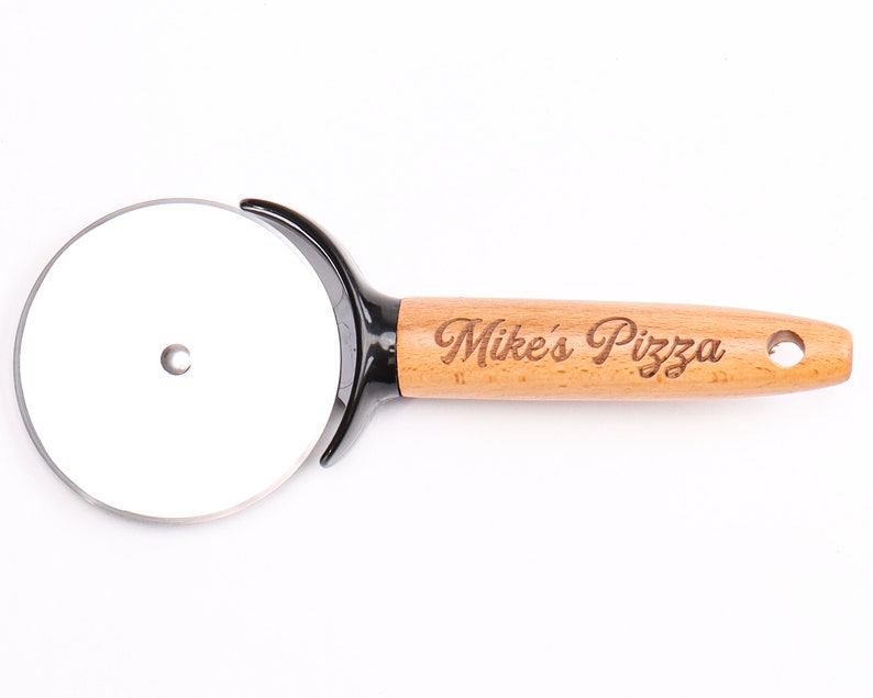 Op de afbeelding: Pizzasnijder met een roestvrijstalen mes en een houten handvat. De handgreep is gegraveerd met de woorden "Mike's Pizza". Het heeft een zwarte plastic bescherming en een gat om op te hangen.