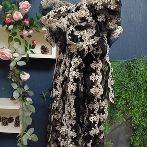 Puede incluir: Un lazo de encaje negro y dorado con un botón dorado grande y decorativo en el centro. El lazo está colgado de una cuerda y está rodeado de vegetación.