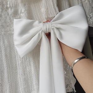 Peut inclure: Un grand nœud en satin blanc avec de longues queues. Le nœud est noué autour d'un tissu en dentelle blanche.