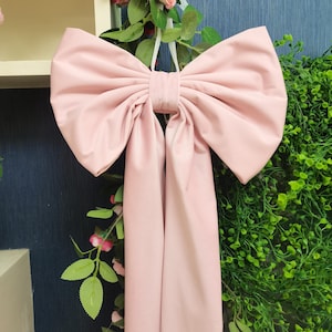 Puede incluir: Un gran lazo de satén rosa claro con colas largas. El lazo está atado con una cinta blanca y cuelga de una guirnalda floral.