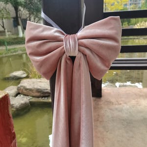 May include: A large, blush pink velvet bow tied around a wooden post. The bow is made of soft, luxurious fabric and has a long, flowing tail.