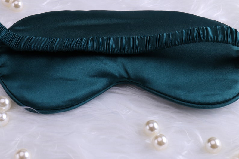 May include: A teal blue silk sleep mask with an elastic band. The mask is on a white fluffy surface.