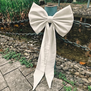 May include: A large, cream-coloured satin bow with long, flowing tails. The bow is tied around a blue metal pole, with a chain fence and water feature in the background. The bow's fabric has a subtle sheen.