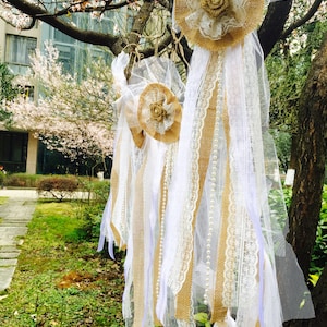 Puede incluir: Decoraciones de boda o evento colgadas de un árbol. Las decoraciones presentan arpillera, encaje y cintas de tela blancas, acentuadas con hilos de perlas y adornos florales. La paleta de colores incluye blanco, beige y dorado.