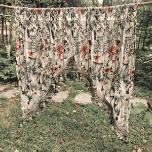 Könnte beinhalten: Ein halbtransparenter, cremeweißer Stoffbehang mit einem detaillierten botanischen Druck aus dunkelgrünem Blattwerk und leuchtend rot-orangen Blüten. Die Unterkante ist bogenförmig geschnitten und mit einer Zierborte sowie hängenden Quasten versehen.