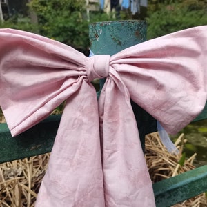Könnte beinhalten: Ein großer rosa Schleife, die um einen grünen Metallzaunpfosten gebunden ist. Die Schleife besteht aus einem weichen, zerknitterten Stoff mit einem dezenten Marmormuster.