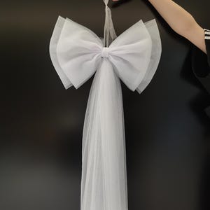 Peut inclure: Un grand nœud en tulle blanc avec une longue queue flottante. Le nœud est suspendu par un ruban transparent. Le nœud est composé de plusieurs couches de tissu doux et transparent. Le fond est de couleur sombre et unie.