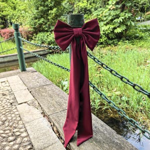 Peut inclure: Un grand nœud en satin bordeaux avec de longues queues flottantes est attaché à un poteau en métal vert. Le nœud est le point central, avec une forme luxuriante et pleine. L'arrière-plan présente une clôture en chaîne verte, une corniche en bois et une verdure luxuriante.