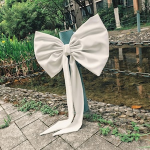 Puede incluir: Un lazo blanco grande de tela, atado alrededor de un poste azul. El lazo tiene largas colas fluidas y se encuentra sobre un fondo de vegetación y agua. El lazo decorativo es ideal para eventos.