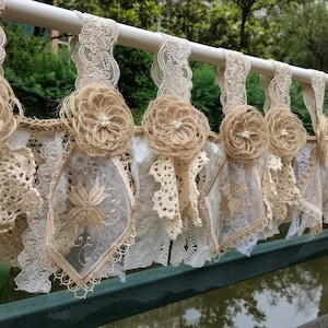 May include: A rustic-style window valance with a beige and white color scheme. It features a row of burlap flowers with pearl accents, and lace trim.