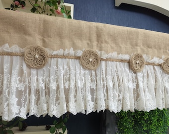 May include: A burlap valance with a white lace trim and burlap rosettes. The valance is a light brown color and the lace is white.