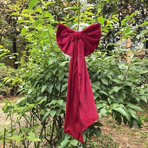 Könnte beinhalten: Eine große, weinrote Samtschleife mit langen, fließenden Enden hängt an einem Ast. Die Schleife hat eine tiefrote Farbe und scheint aus einem weichen, luxuriösen Stoff zu bestehen. Der Hintergrund ist mit grünem Laub gefüllt.
