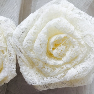Puede incluir: Dos adornos florales de encaje blanco, cada uno con un diseño delicado y en capas. Las flores están hechas de encaje blanco y tienen un aspecto suave y romántico.