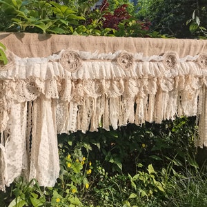May include: A rustic burlap valance with a layer of delicate white lace and fabric flowers. The valance is trimmed with a fringe of lace and fabric.