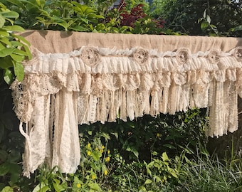 May include: A rustic burlap valance with a layer of delicate white lace and fabric flowers. The valance is trimmed with a fringe of lace and fabric.