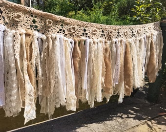 May include: A rustic-style garland with a crocheted top and long, flowing fabric strands in shades of white and beige.