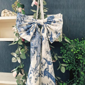 Puede incluir: Un gran lazo decorativo hecho de tela blanca con un estampado azul toile de Jouy que presenta pájaros y elementos arquitectónicos. El lazo está suspendido por una cinta blanca contra una pared azul oscuro, con vegetación y flores alrededor.