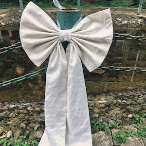 May include: A large, off-white fabric bow tied around a teal metal pole. The bow has long, flowing tails and is set against a backdrop of a chain fence and a stone wall. The bow is likely used for decoration.