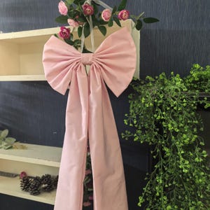 May include: A large, light pink bow with long, flowing tails hangs against a dark blue wall. The bow is made of a smooth fabric and is adorned with pink roses and greenery, suggesting a decorative or event-themed item.